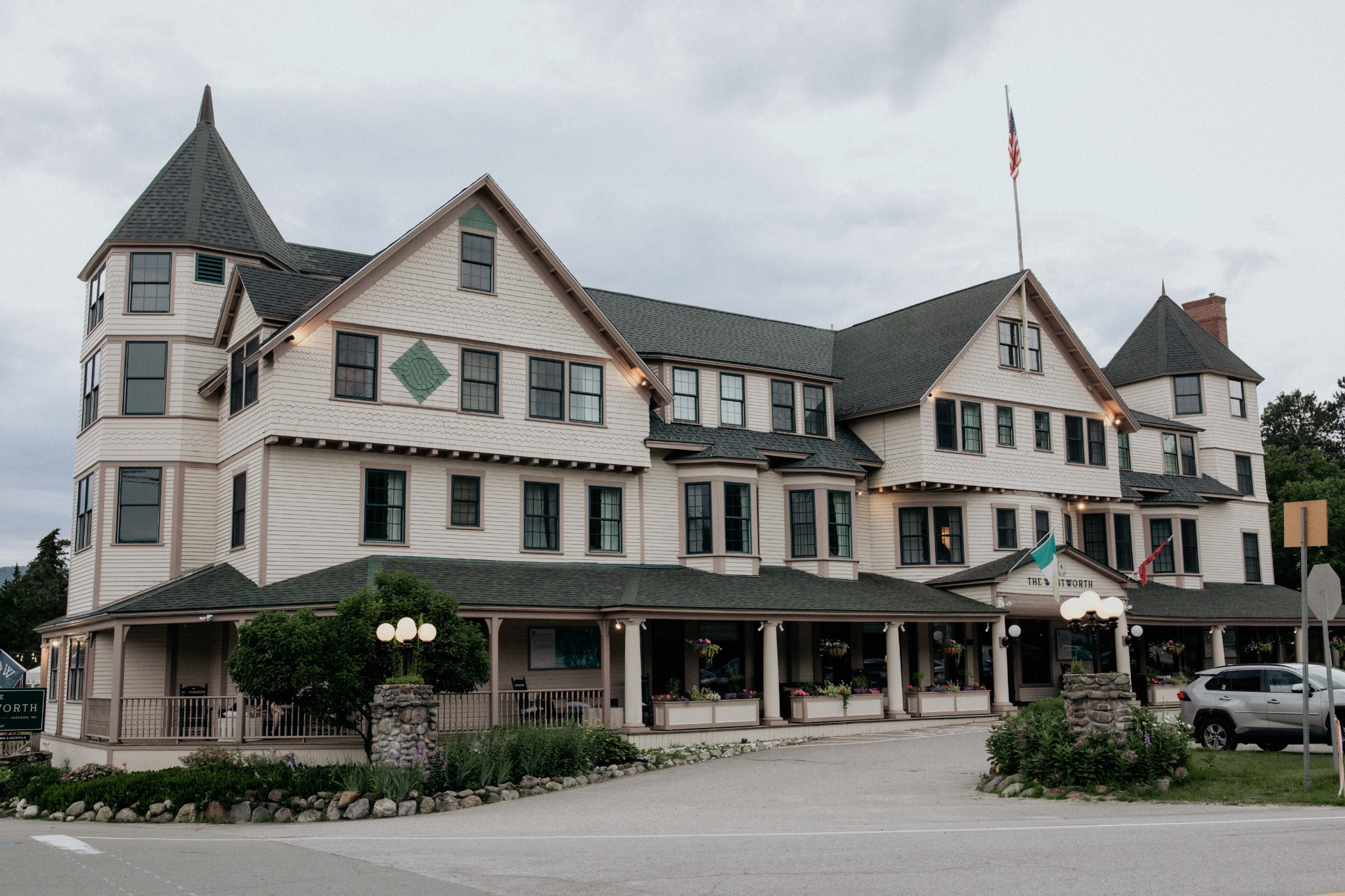 A large, three-story building with a gray roof and white siding, featuring a covered porch and a car parked in front.