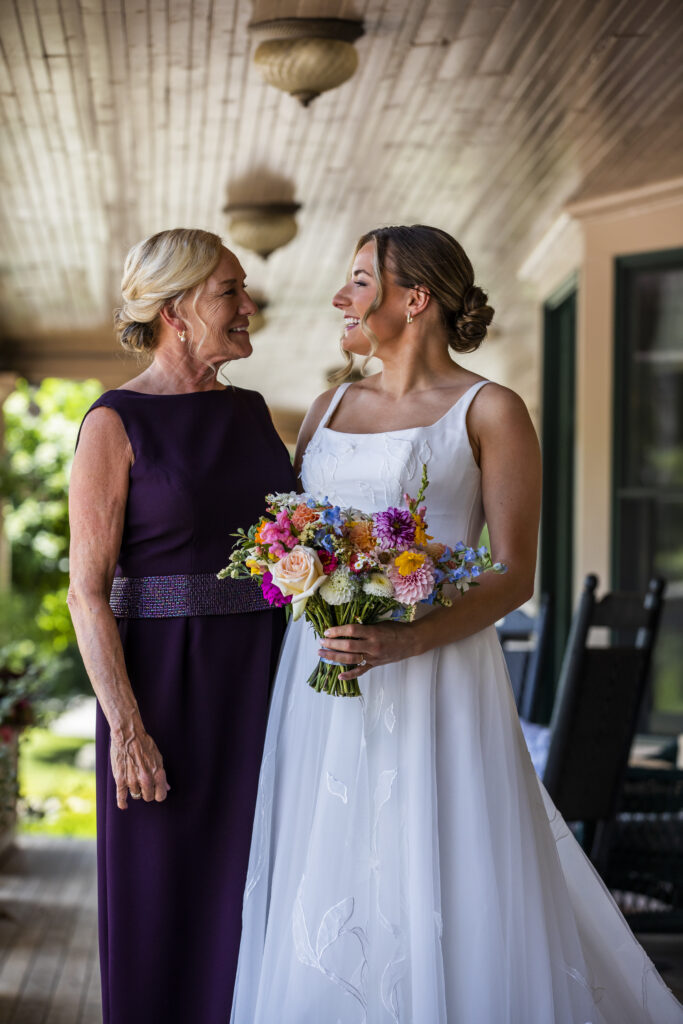 Two women in formal attire, one holding a bouquet of flowers. The woman on the left has blonde hair and is wearing a purple dress, while the woman on the right has brown hair and is wearing a white dress.