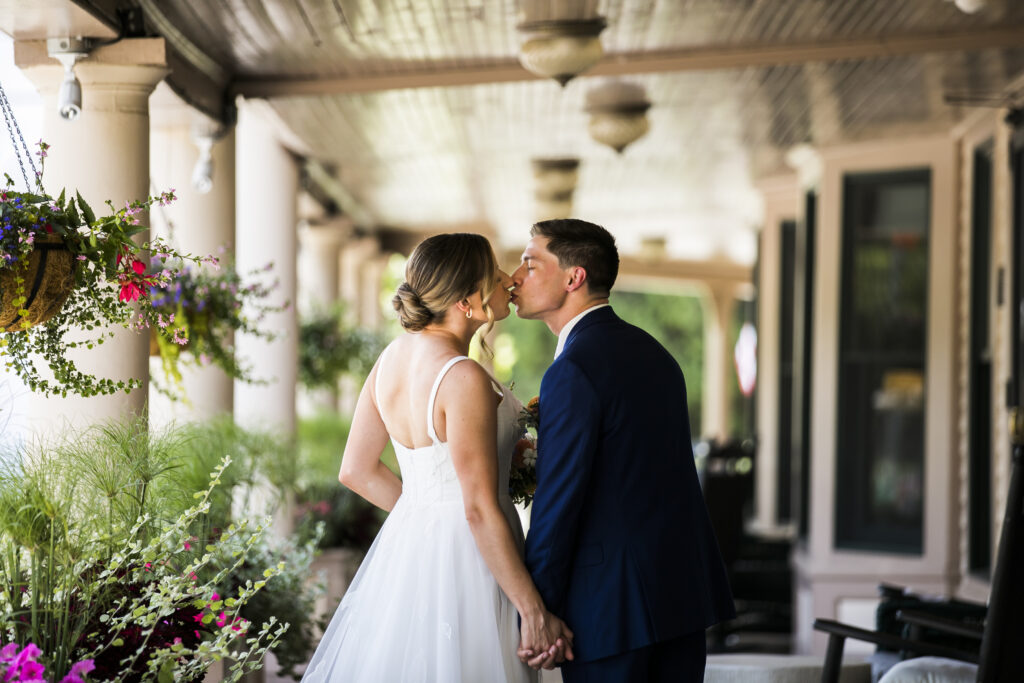 A bride and groom standing in a covered walkway, holding hands and kissing. The woman is wearing a white wedding dress, and the man is dressed in a dark suit. The background features a row of columns, a doorway, and hanging flowers.