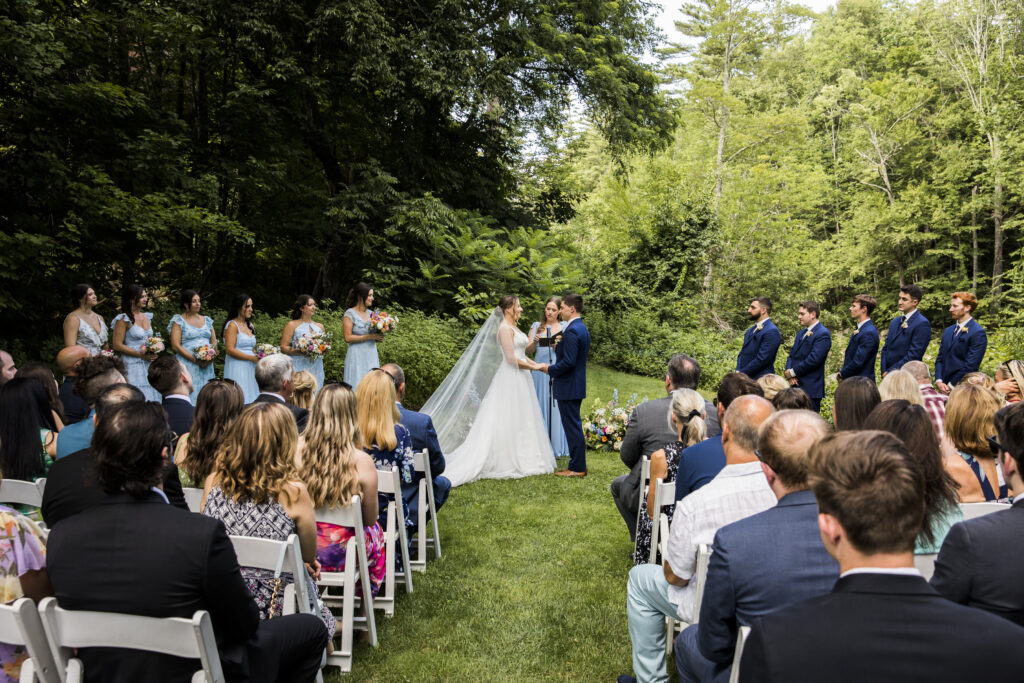A bride and groom standing in a grassy area surrounded by trees, with a group of people seated in chairs watching them.