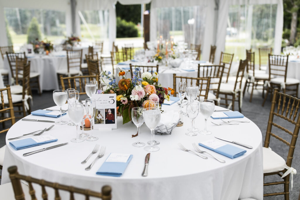 A round table with a white tablecloth, set with silverware, glasses, and a floral centerpiece, surrounded by chairs, in a wedding reception setting.