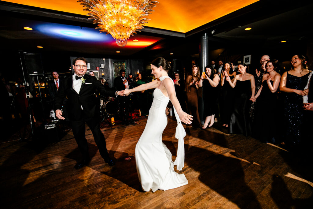 A bride and groom dancing in a dimly lit room with guests watching.