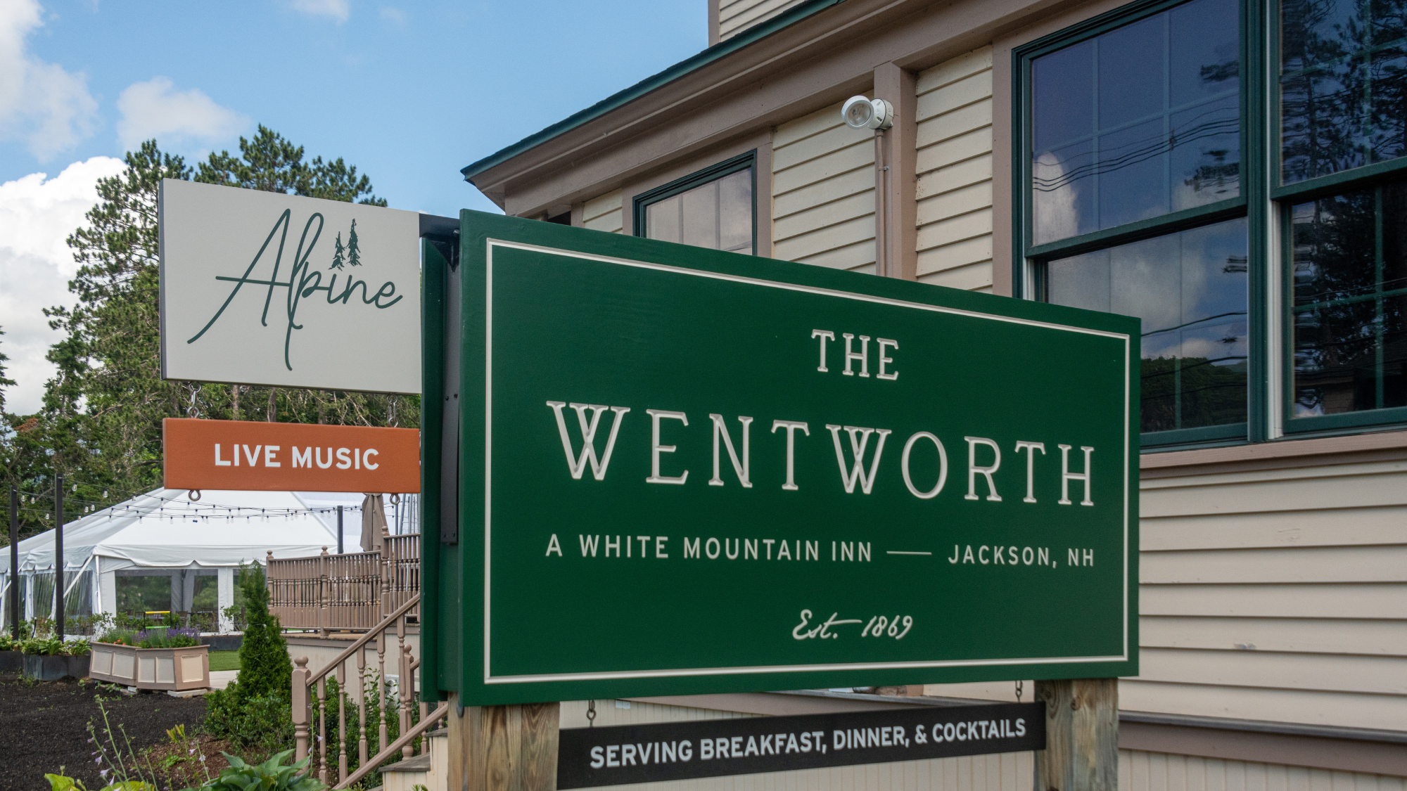 A green sign with white text reading "THE WENTWORTH A WHITE MOUNTAIN INN JACKSON, NH" in front of a building. The sign also includes smaller text at the bottom that reads "SERVING BREAKFAST, DINNER, & COCKTAILS".