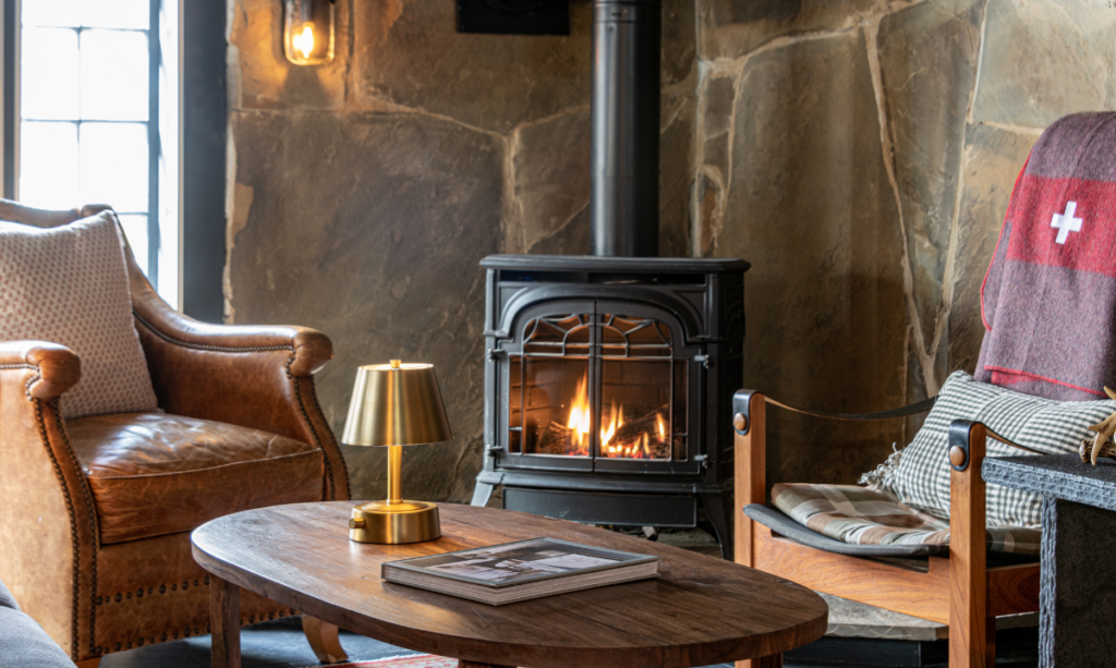 A cozy living room scene with a stone wall, wood-burning stove, and comfortable seating. The room features a dark wood coffee table, a leather armchair, a wooden chair, and a lamp. The atmosphere is warm and inviting.